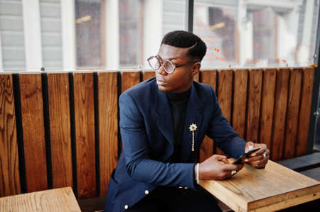 Amazingly looking african american man wear at blue blazer with brooch, black turtleneck and glasses posed at street. Fashionable black guy sitting behind table with mobile phone.の写真素材