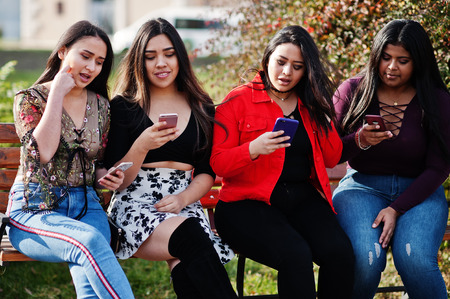 Group of four happy and pretty latino girls from Ecuador posed at street and looking at mobile...