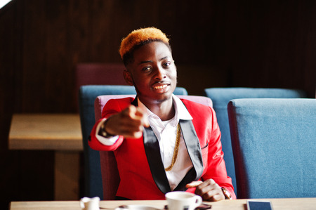 Fashion african american man model at red suit, with highlights hair sitting at cafe with coffee and show finger to you.の写真素材