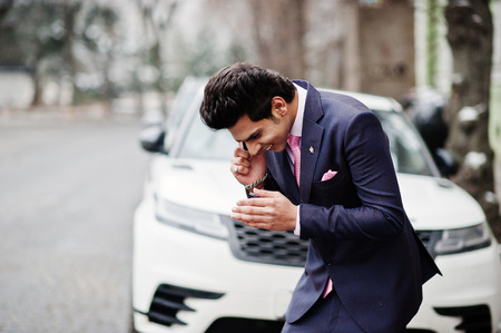 Elegant indian macho man model on suit and pink tie posed against white business car and speaking on...