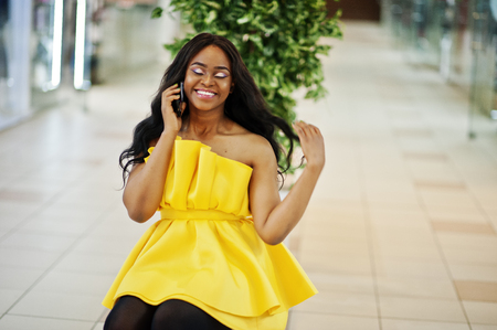 Stylish african american woman at yellow dreess posed at mall and sitting on bench inside, speaking at phone.の写真素材