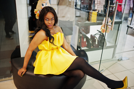 Stylish african american woman at yellow dreess sitting on a puff against showcases at mall.の写真素材