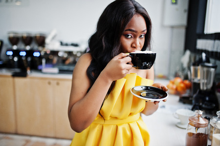 Stylish african american woman at yellow dreess posed at coffee house with cup of cappuccino.