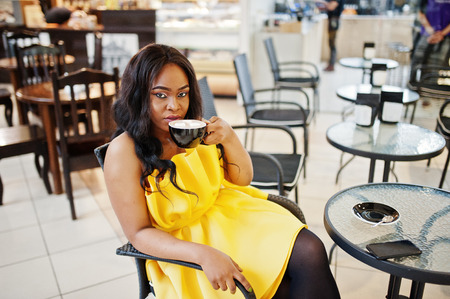 Stylish african american woman at yellow dreess posed at cafe with cup of cappuccino.