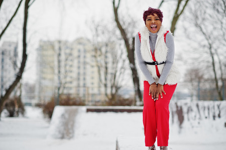 African american woman in red pants and white fur coat jacket posed at winter day against snowy background.の写真素材