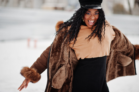 African american woman in sheepskin coat and cap posed at winter day against snowy background.の写真素材