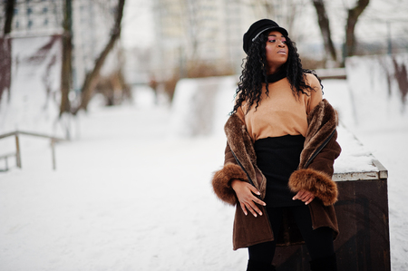 African american woman in sheepskin coat and cap posed at winter day against snowy background.の写真素材