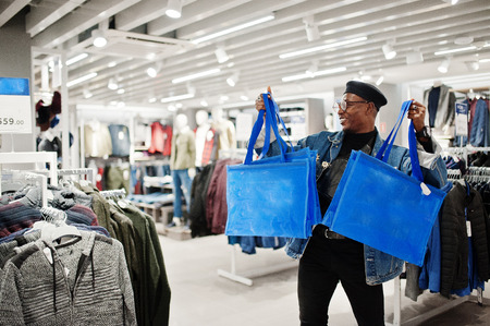 Stylish casual african american man at jeans jacket and black beret with fanny pack or waist bag holding blue shopping bags at clothes store.の写真素材