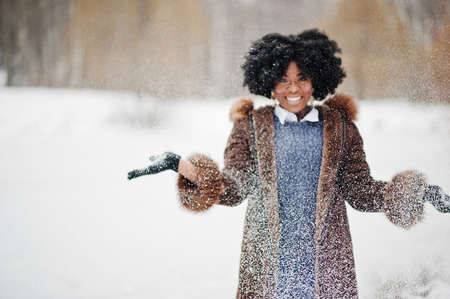 Curly hair african american woman wear on sheepskin coat and gloves posed at winter day throws up snow.の写真素材