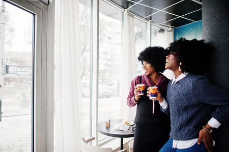 Two curly hair african american woman wear on sweaters with cups of tea posed at cafe indoor.の写真素材