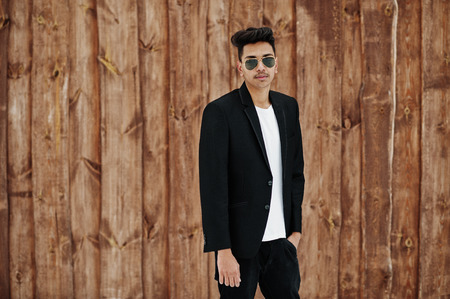 Casual young indian man in black blazer and sunglasses posed against wooden background.の写真素材