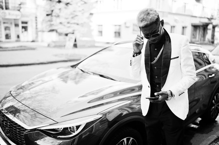 Chic handsome african american man in white suit against red luxury car with mobile phone at hand.の写真素材