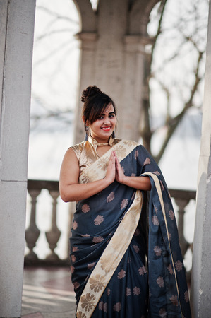 Elegant brunette south asian indian girl in saree standing under arch and show namaste hands sign.