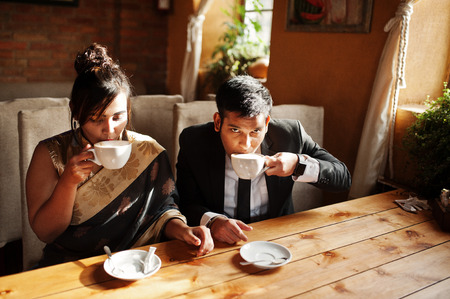 Elegant and fashionable indian friends couple of woman in saree and man in suit sitting on cafe and drinking cappuccino.の写真素材