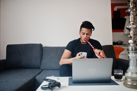 Handsome and fashionable indian man in black sitting at room, smoking hookah, looking on phone with laptop.の写真素材
