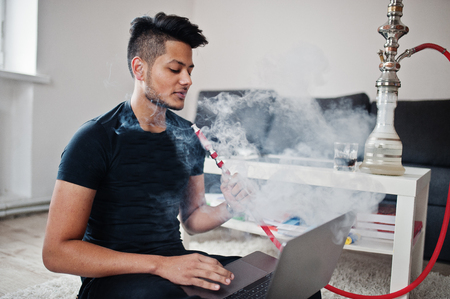 Handsome and fashionable indian man in black sitting at room, smoking hookah and working on laptop.の写真素材