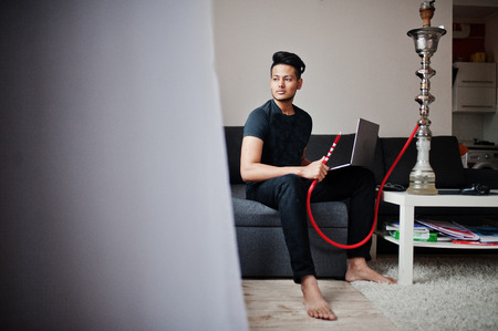 Handsome and fashionable indian man in black sitting at room, smoking hookah and working on laptop.の写真素材