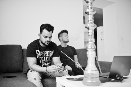 Two handsome and fashionable indian mans relaxing at room and smoking hookah. One of them looking at mobile phone.の写真素材