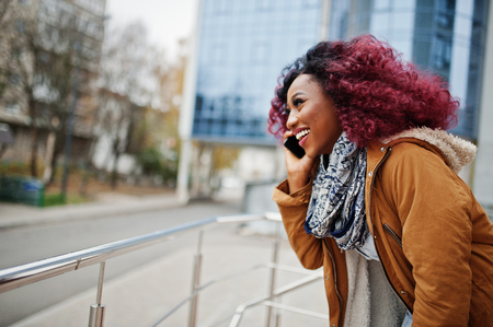 Attractive curly african american woman in brown coat posed near railings against modern multistory building speaking on mobile phone.の写真素材