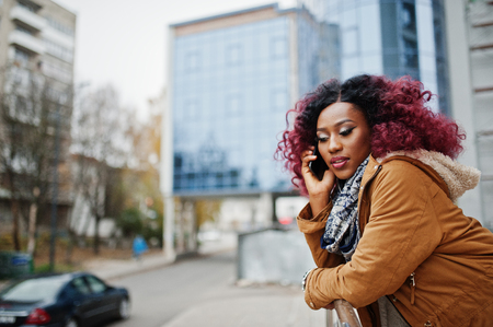 Attractive curly african american woman in brown coat posed near railings against modern multistory building speaking on mobile phone.の写真素材