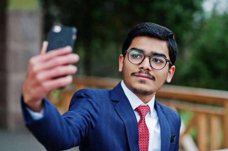 Indian young man at glasses, wear on suit with red tie posed outdoor and making selfie on mobile phone.の写真素材