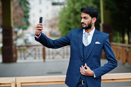 Stylish beard indian business man with bindi on forehead, wear on blue suit posed outdoor and making selfie at mobile phone.の写真素材