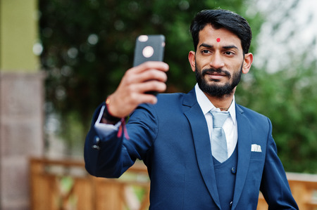 Stylish beard indian business man with bindi on forehead, wear on blue suit posed outdoor and making selfie at mobile phone.の写真素材