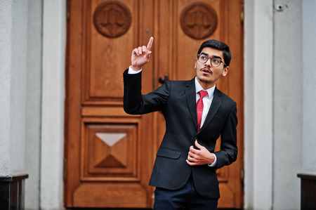 Indian young man at glasses, wear on black suit with red tie posed outdoor against door of building show finger up, he has idea.の写真素材