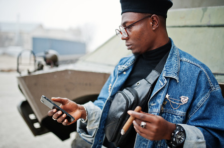 African american man in jeans jacket, beret and eyeglasses, smoking cigar and posed against btr military armored vehicle, with mobile phone at hand.の写真素材