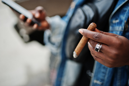 Close up hands of african american man in jeans jacket,  smoking cigar with mobile phone at hand.の写真素材