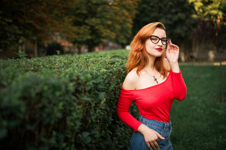 Attractive redhaired woman in eyeglasses, wear on red blouse and jeans skirt posing at green park.の写真素材