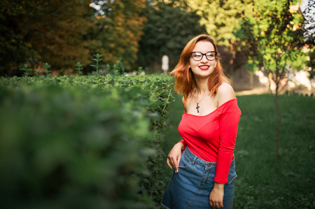 Attractive redhaired woman in eyeglasses, wear on red blouse and jeans skirt posing at green park.の写真素材