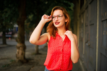 Attractive redhaired woman in eyeglasses, wear on red blouse and jeans skirt posing.の写真素材