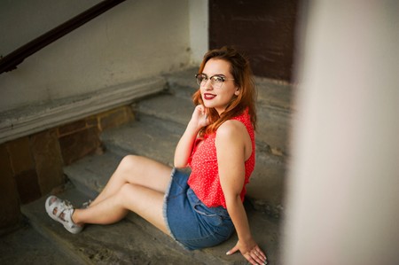 Attractive redhaired woman in eyeglasses, wear on red blouse and jeans skirt posing.の写真素材