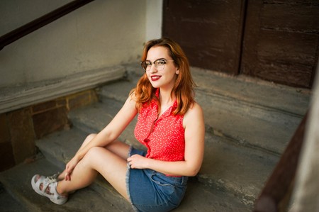 Attractive redhaired woman in eyeglasses, wear on red blouse and jeans skirt posing.の写真素材
