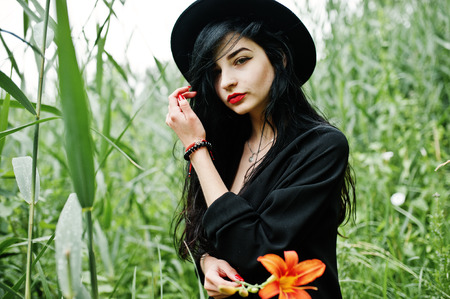Sensual girl all in black, red lips and hat. Goth dramatic woman hold orange lily flower on  common reed.の写真素材