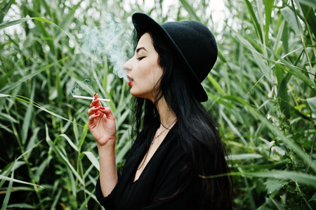 Sensual smoker girl all in black, red lips and hat. Goth dramatic woman smoking on common reed.の写真素材