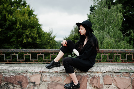 Sensual girl all in black, red lips and hat. Goth dramatic woman hold white chrysanthemum flower.の写真素材