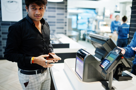 Indian man holding wallet behind his pay cash desk with order screen and card payment terminal in...