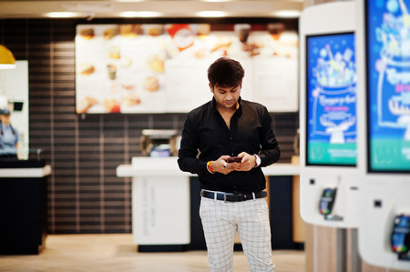 Stylish indian man posed at fast food cafe near self pay floor kiosk with mobile phone at hand.