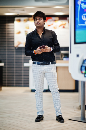 Stylish indian man posed at fast food cafe near self pay floor kiosk with mobile phone at hand.