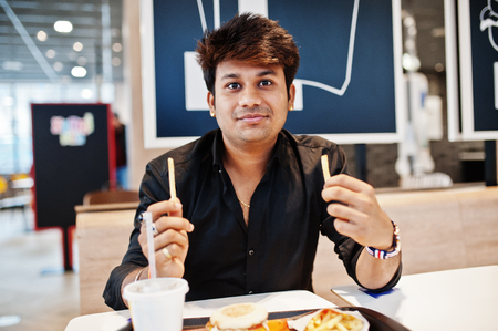 Stylish indian man sitting at fast food cafe and eating french fries.の写真素材