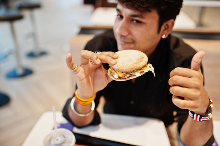 Stylish indian man sitting at fast food cafe and eating hamburger. Show thumb up.の写真素材