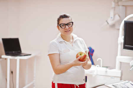 Portrait of female dentist woman at glasses standing in her dentistry office with an apple and toothbrush.の写真素材