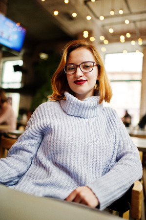 Cheerful young beautiful redhaired woman in glasses, warm wool sweater sitting at her working place on cafe.の写真素材