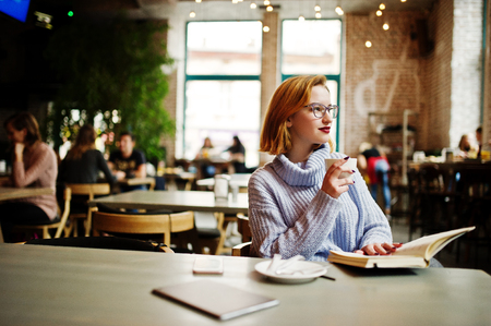 Cheerful young beautiful redhaired woman in glasses sitting at her working place on cafe and drinking coffee.の写真素材