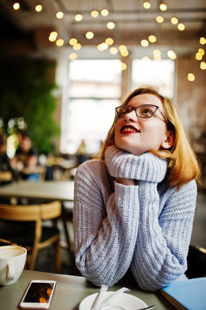 Cheerful young beautiful redhaired woman in glasses, warm wool sweater sitting at her working place on cafe.の写真素材
