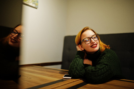Cheerful young beautiful redhaired woman in glasses, wear on green warm wool sweater,  sitting at...