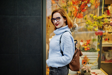 Cheerful young beautiful redhaired woman in glasses, warm blue wool sweater with backpack posed outdoor against autumn leaves tree decorations on store.の写真素材