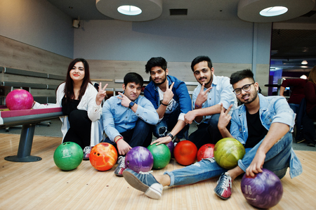 Group of five south asian peoples having rest and fun at bowling club. Holding bowling balls, sitting on alley and show two fingers sign.の写真素材